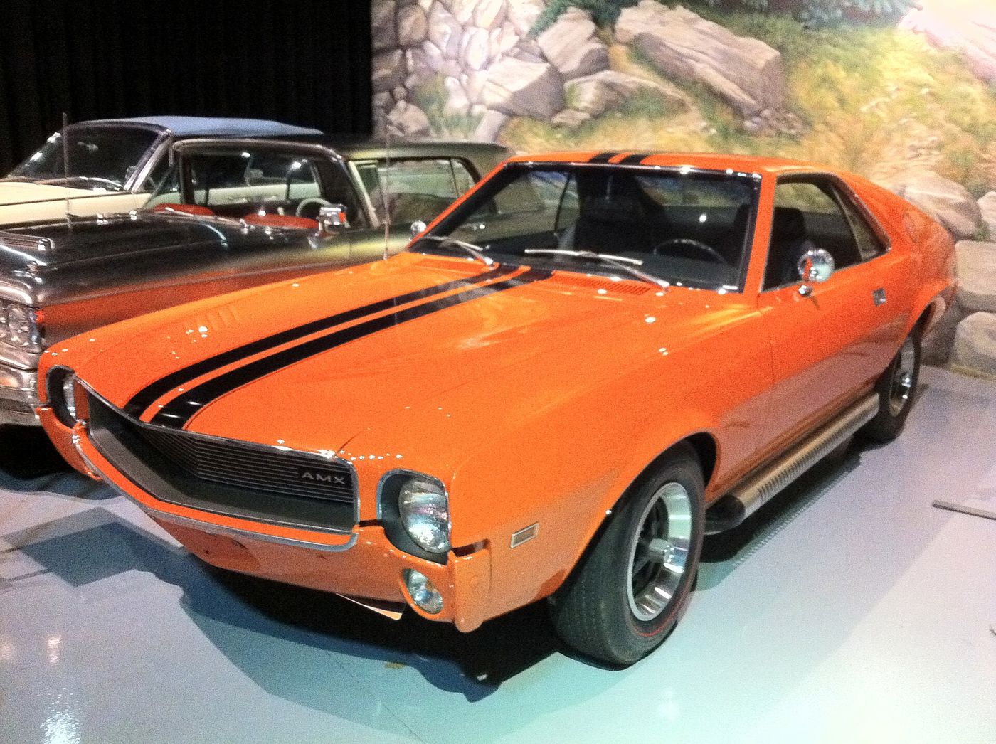 Orange 1969 AMC AMX two-seater from the front three-quarter, with black over-the-top stripes, AMX grille badge and side exhaust trim, displayed indoors