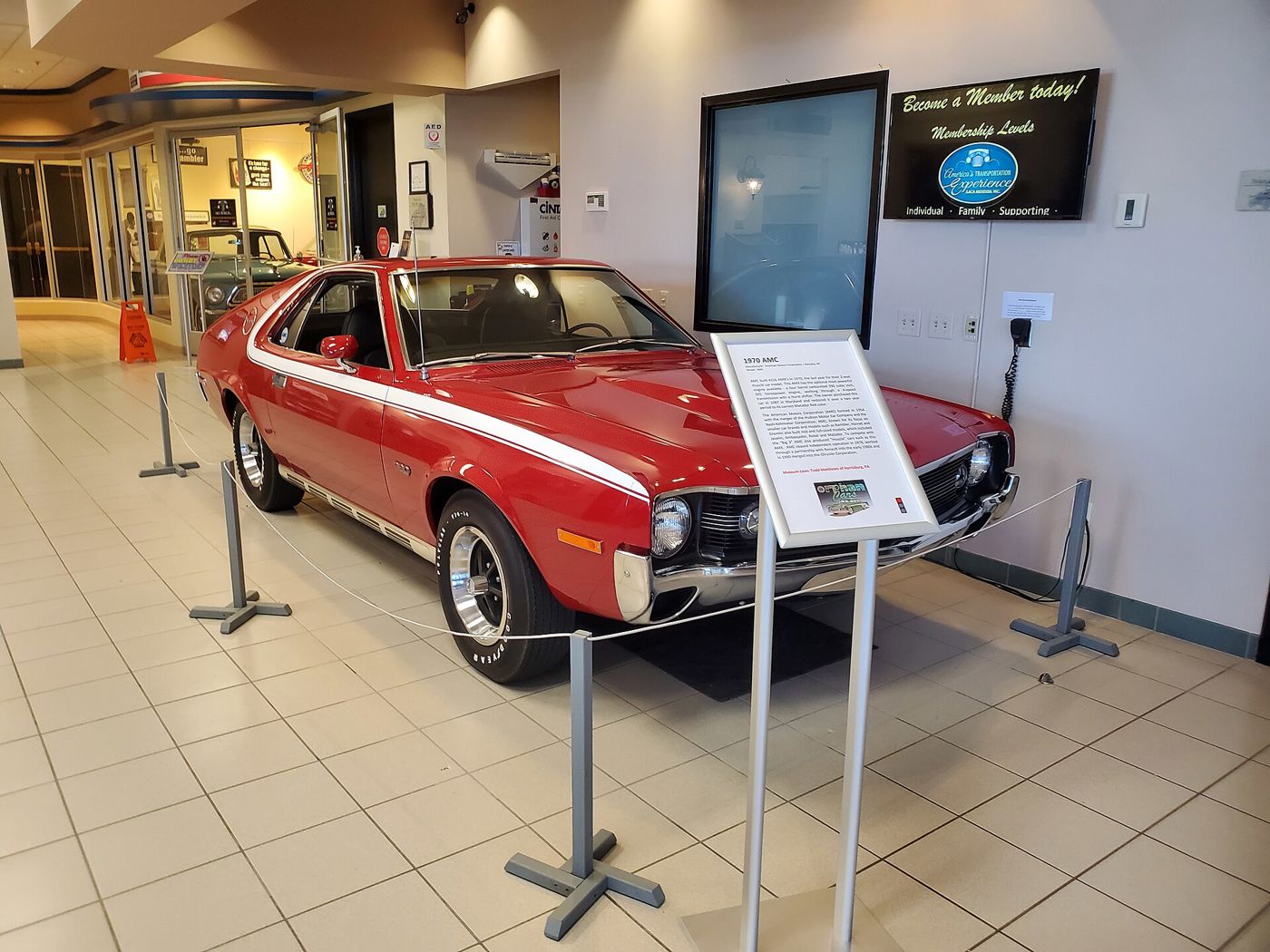Red 1970 AMC AMX with a white C-stripe, 390 fender badge and 1970 grille, on display behind stanchions in a museum lobby