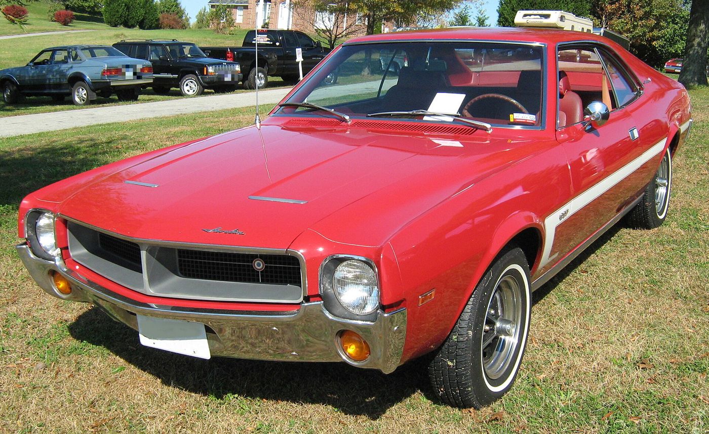 Red 1969 AMC Javelin SST hardtop from the front three-quarter on grass, showing the 1969 grille with its centre bull's-eye emblem, twin hood vents and a white C-shaped side stripe