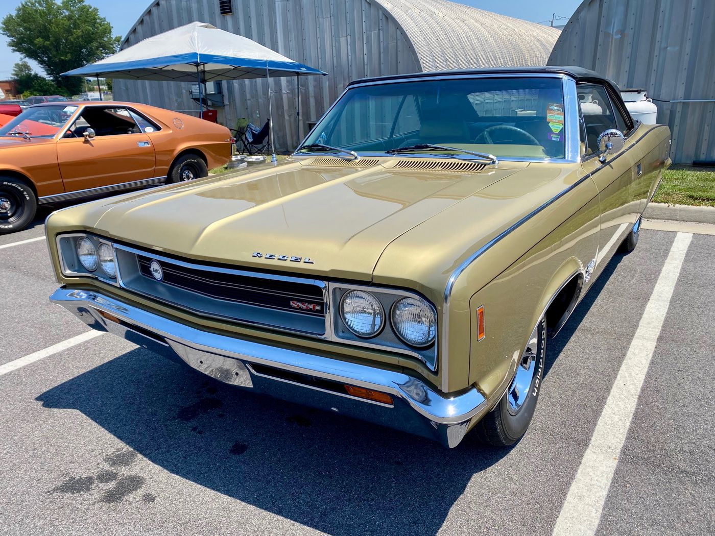 Front three-quarter view of a gold 1968 AMC Rebel SST convertible with a black top, with an orange AMX partly visible at left