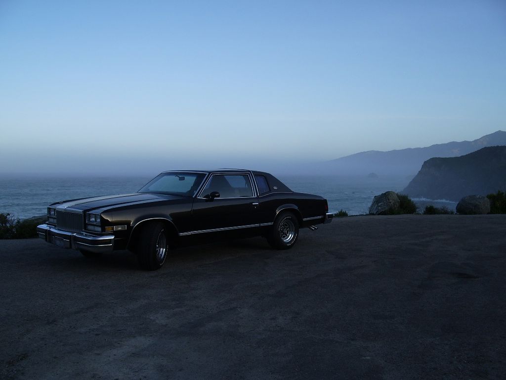 Dark 1977 Buick Riviera coupe silhouetted at dusk on a coastal overlook