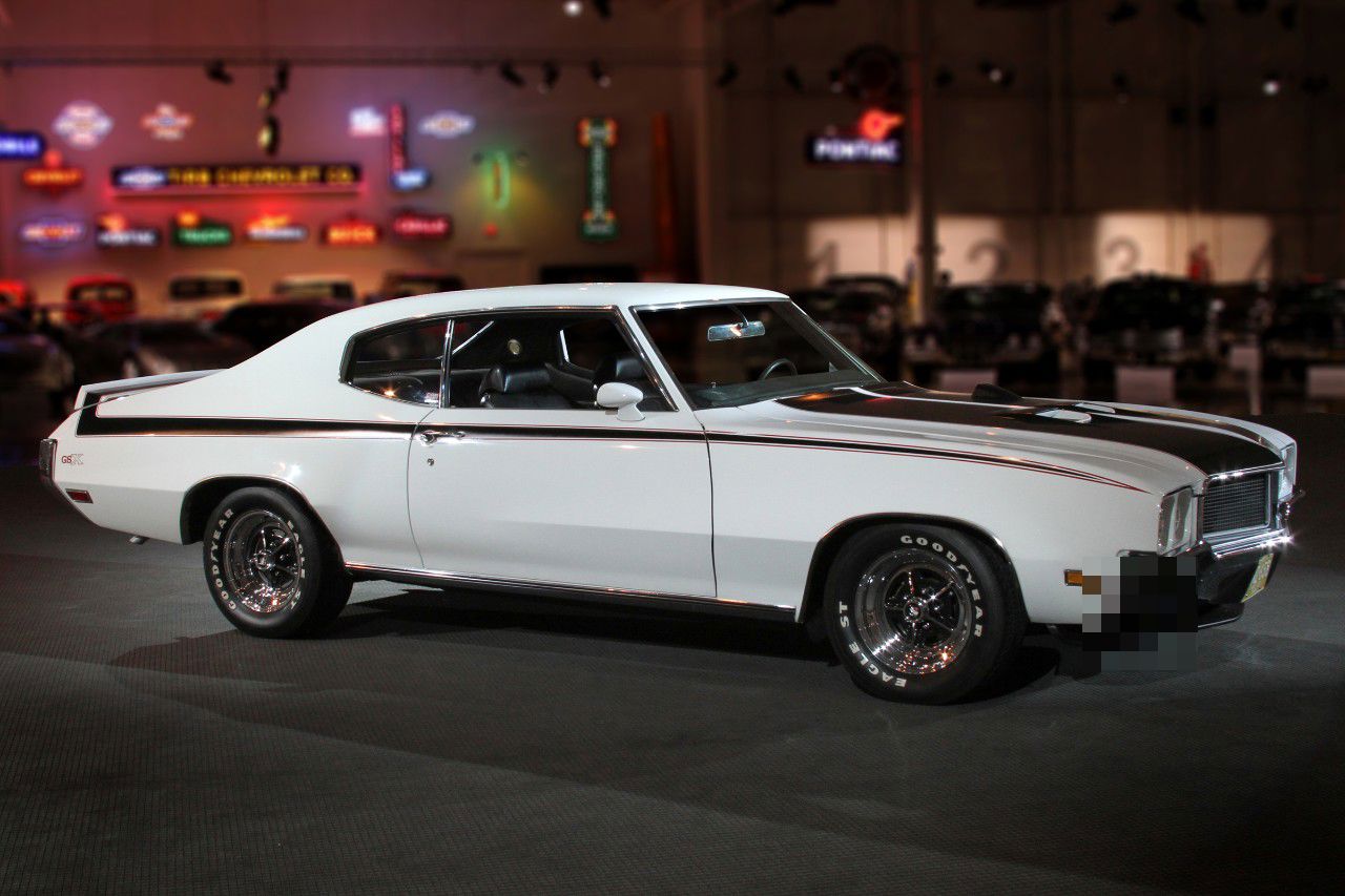 White 1970 Buick GSX with black stripes, hood scoops and rear spoiler, front three-quarter view in a museum hall