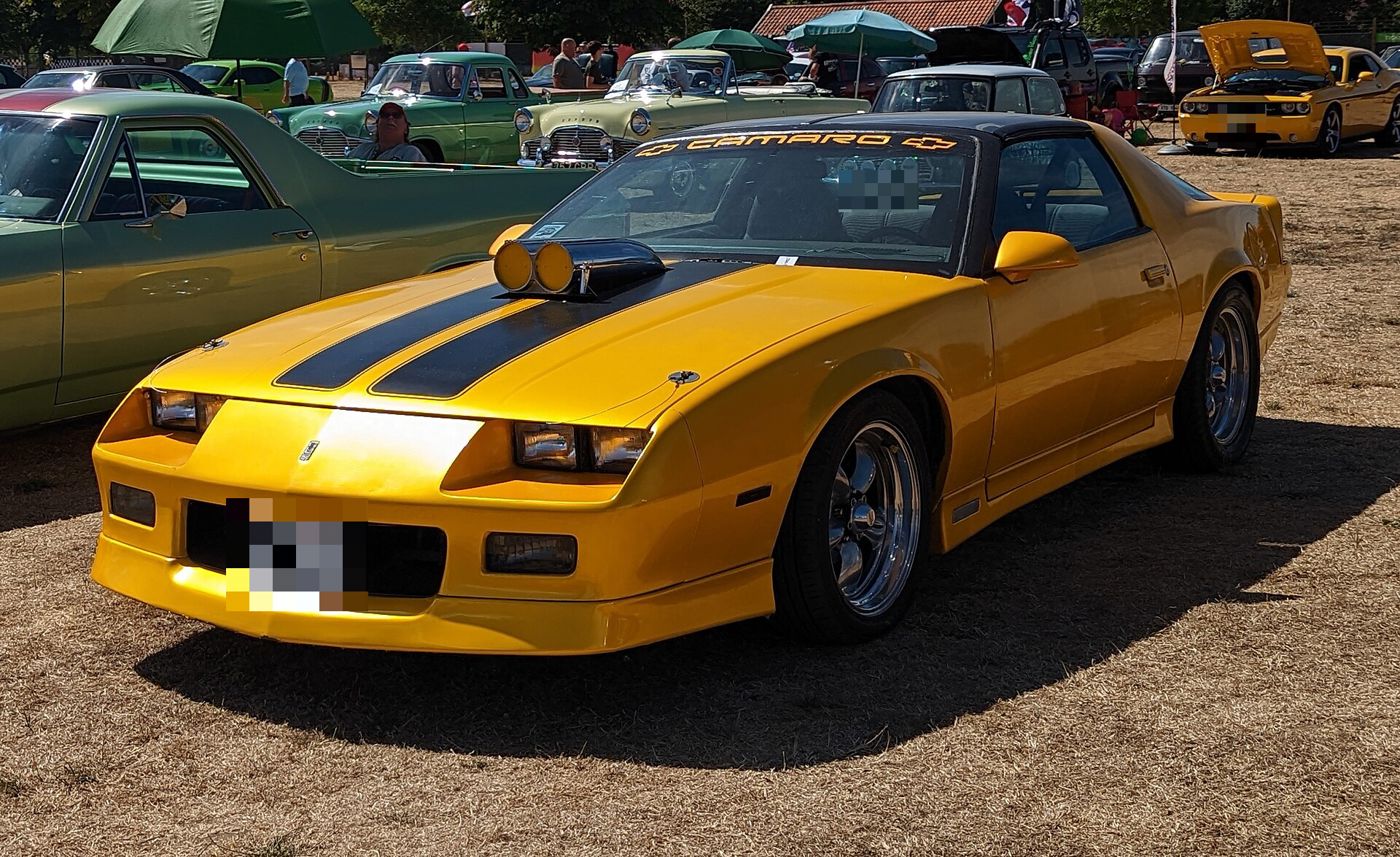 Yellow third-generation 1987 Chevrolet Camaro with black hood stripes, a non-factory twin-inlet hood scoop and aftermarket wheels, at a British car show