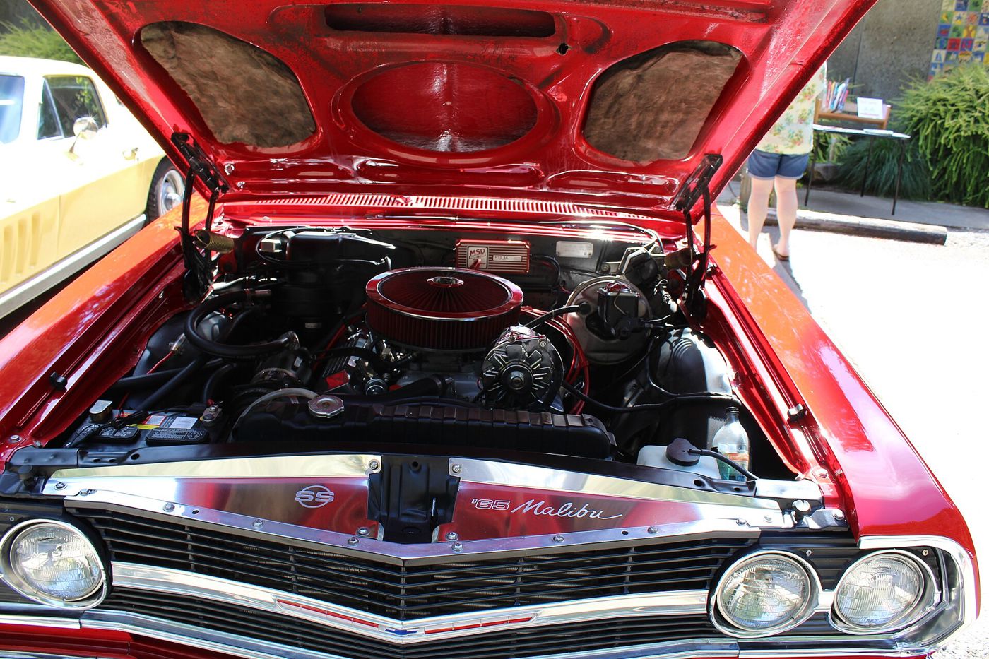 Engine bay of a red 1965 Chevrolet Chevelle Malibu SS with a chrome open-element air cleaner, MSD ignition box and a polished radiator panel reading SS and 65 Malibu