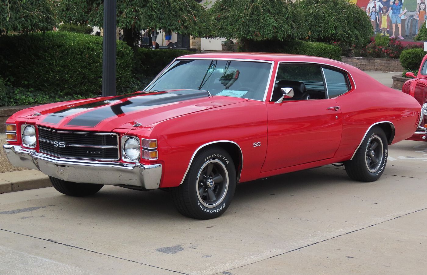 Red 1971 Chevrolet Chevelle SS two-door hardtop at a downtown car show, front three-quarter, with the single-headlamp 1971 front end, SS grille and fender badges, black hood stripes and hood pins
