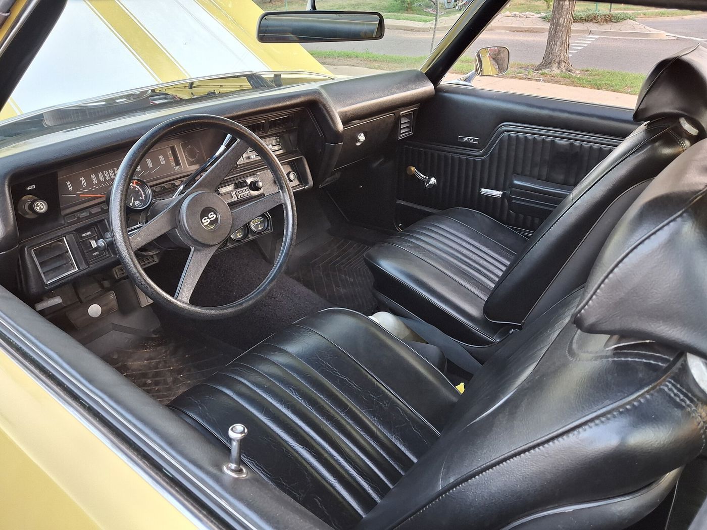 Black interior of a yellow 1972 Chevrolet Chevelle SS 454 showing black bucket seats, the SS steering wheel, SS door-panel badge, horizontal speedometer, auxiliary gauges under the dash and an aftermarket cassette radio