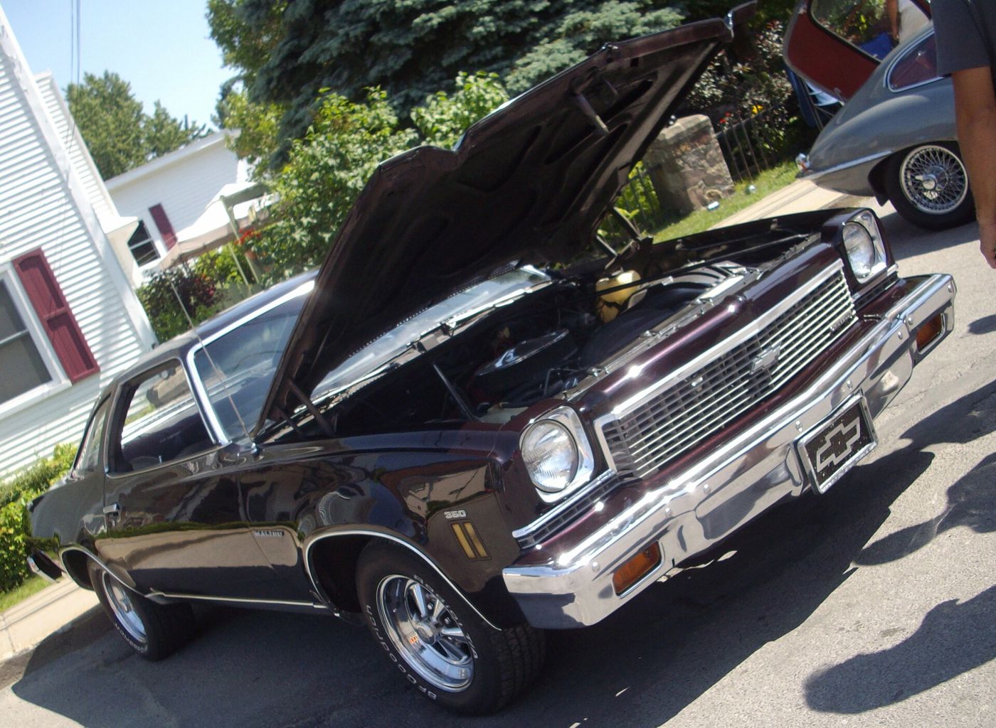 Dark maroon Chevrolet Chevelle Malibu Colonnade hardtop coupe at a street car show, low front three-quarter with the hood raised, showing the single headlamps, bow-tie grille, Malibu fender script and aftermarket wheels