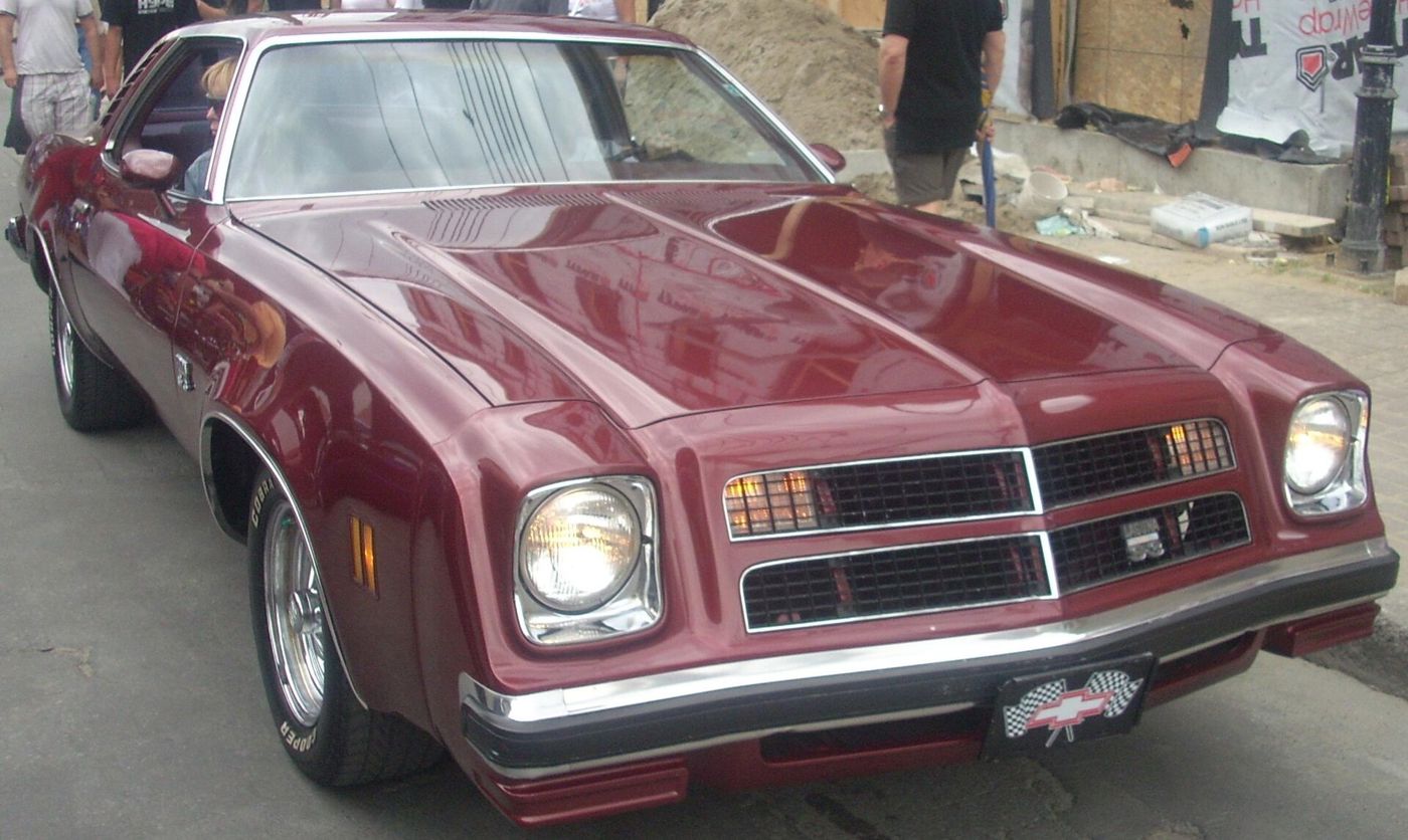 Red 1976 Chevrolet Chevelle Laguna Type S-3 coupe at a street car show, low front three-quarter, showing the 1976 twin-rectangle grille with Laguna badge, body-colour nose and single round headlamps