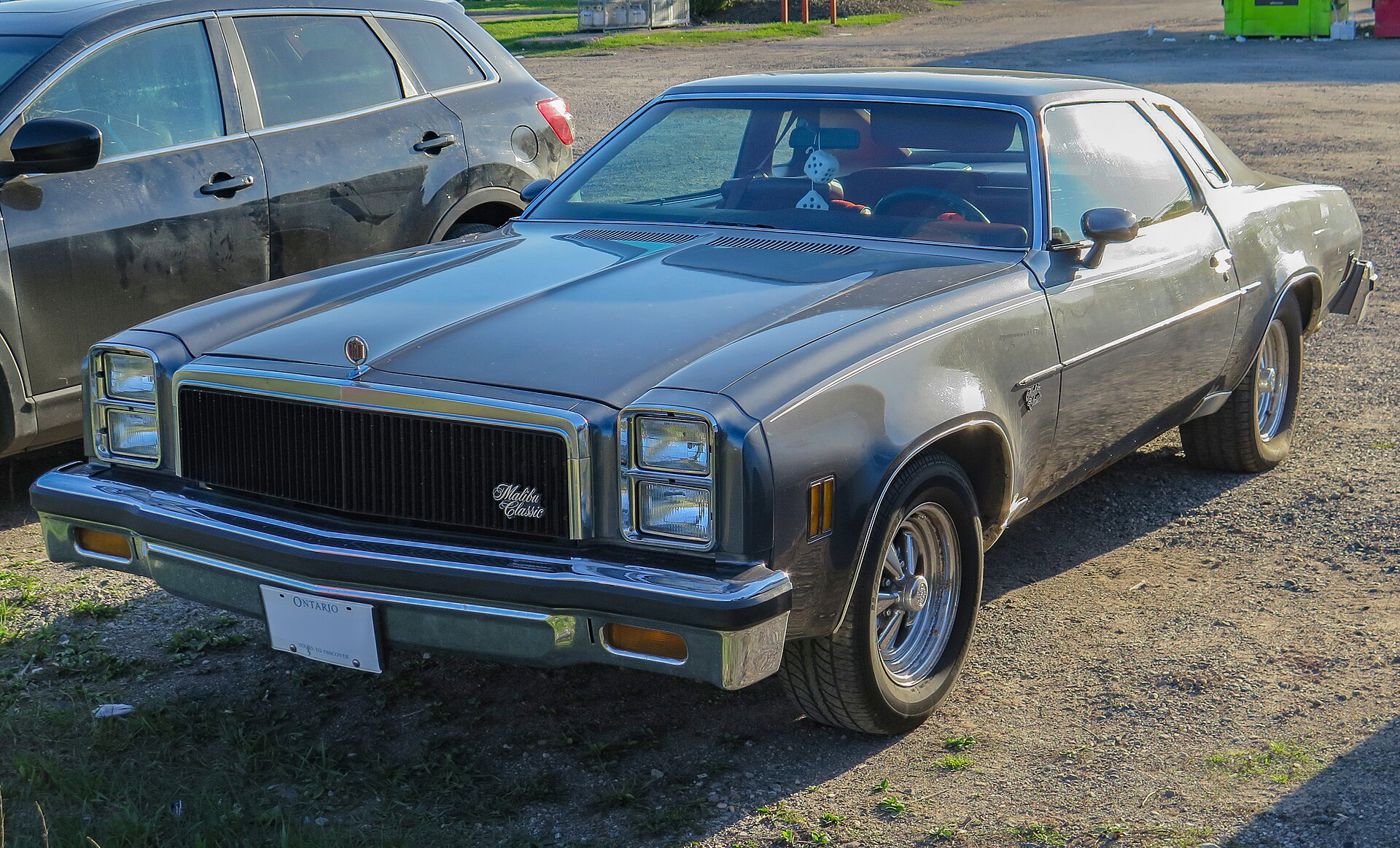Medium gray 1977 Chevrolet Chevelle Malibu Classic Colonnade coupe in a gravel lot, front three-quarter, showing the stacked rectangular headlamps and vertical-bar grille with Malibu Classic script