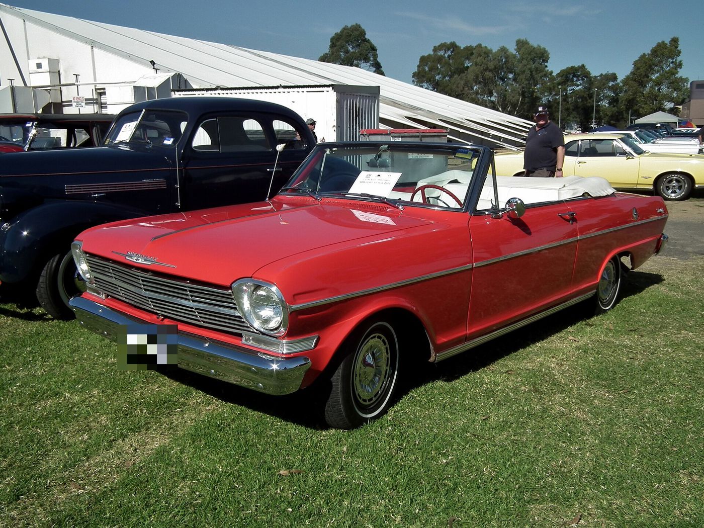 Red 1962 Chevrolet Chevy II Nova 400 convertible with white interior and top down, parked on grass at an Australian car show, front three-quarter, showing the first-year grille and single headlamps