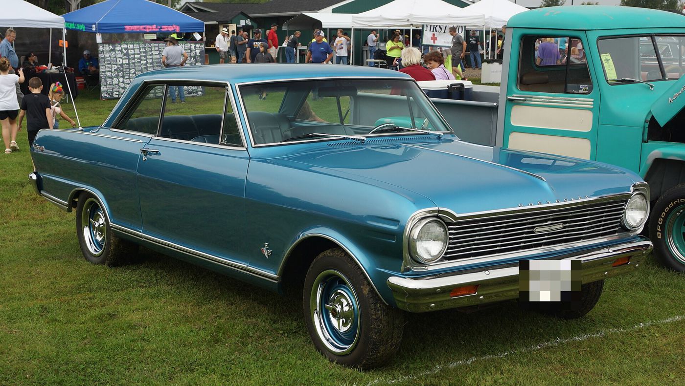 Metallic blue 1965 Chevrolet Chevy II Nova SS two-door hardtop on grass at a car show, front three-quarter, showing the 1965 grille, crossed-flags V8 fender emblem and SS wheel covers