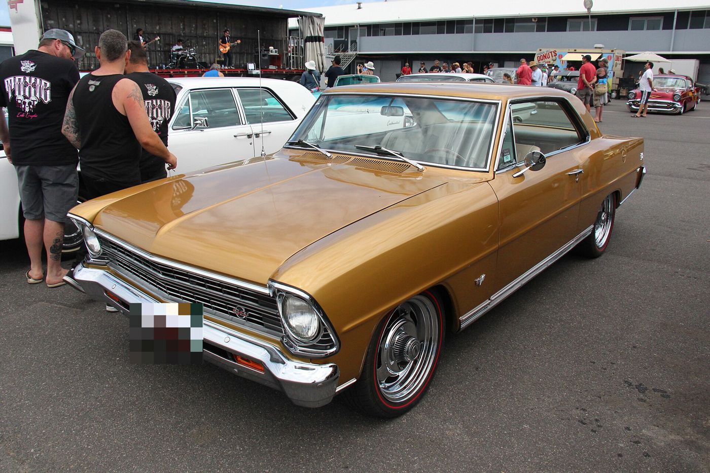 Gold 1967 Chevrolet Chevy II Nova SS two-door hardtop at an Australian car show, front three-quarter, with the SS grille badge and crossed-flags fender emblem; a band plays on a stage behind