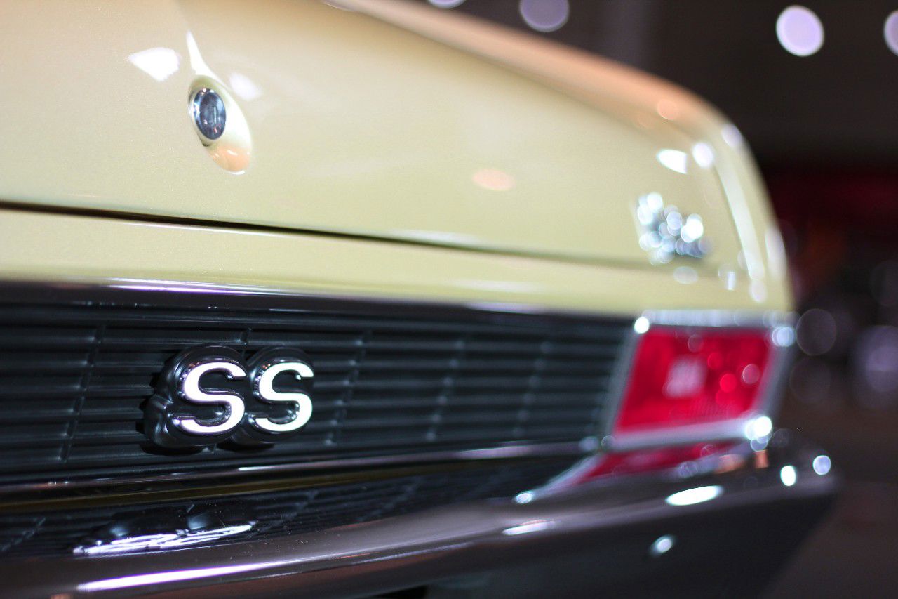 Close-up of the black rear panel of a gold 1970 Chevrolet Nova SS with the chrome SS emblem, the trunk lock and the edge of a taillamp