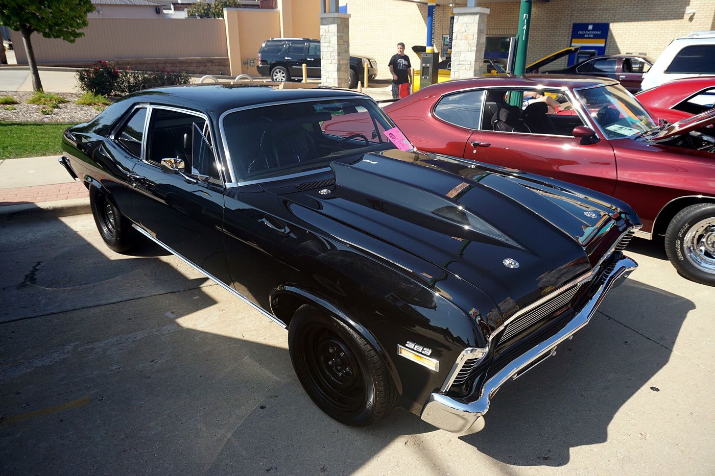 Black 1972 Chevrolet Nova coupe at a downtown car show, front three-quarter, with a raised cowl-induction hood, 383 fender badges and black steel wheels