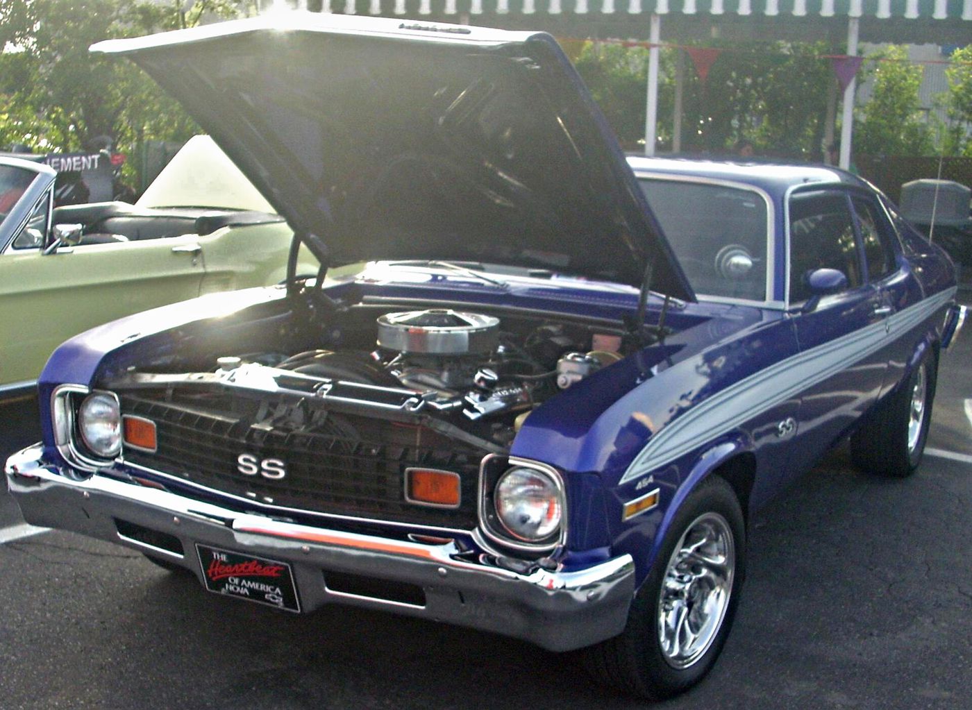 Blue 1973 Chevrolet Nova SS coupe with white side stripes at an evening car show, front three-quarter with the hood raised over a chrome-dressed V8, showing the 1973 SS grille and SS fender badge