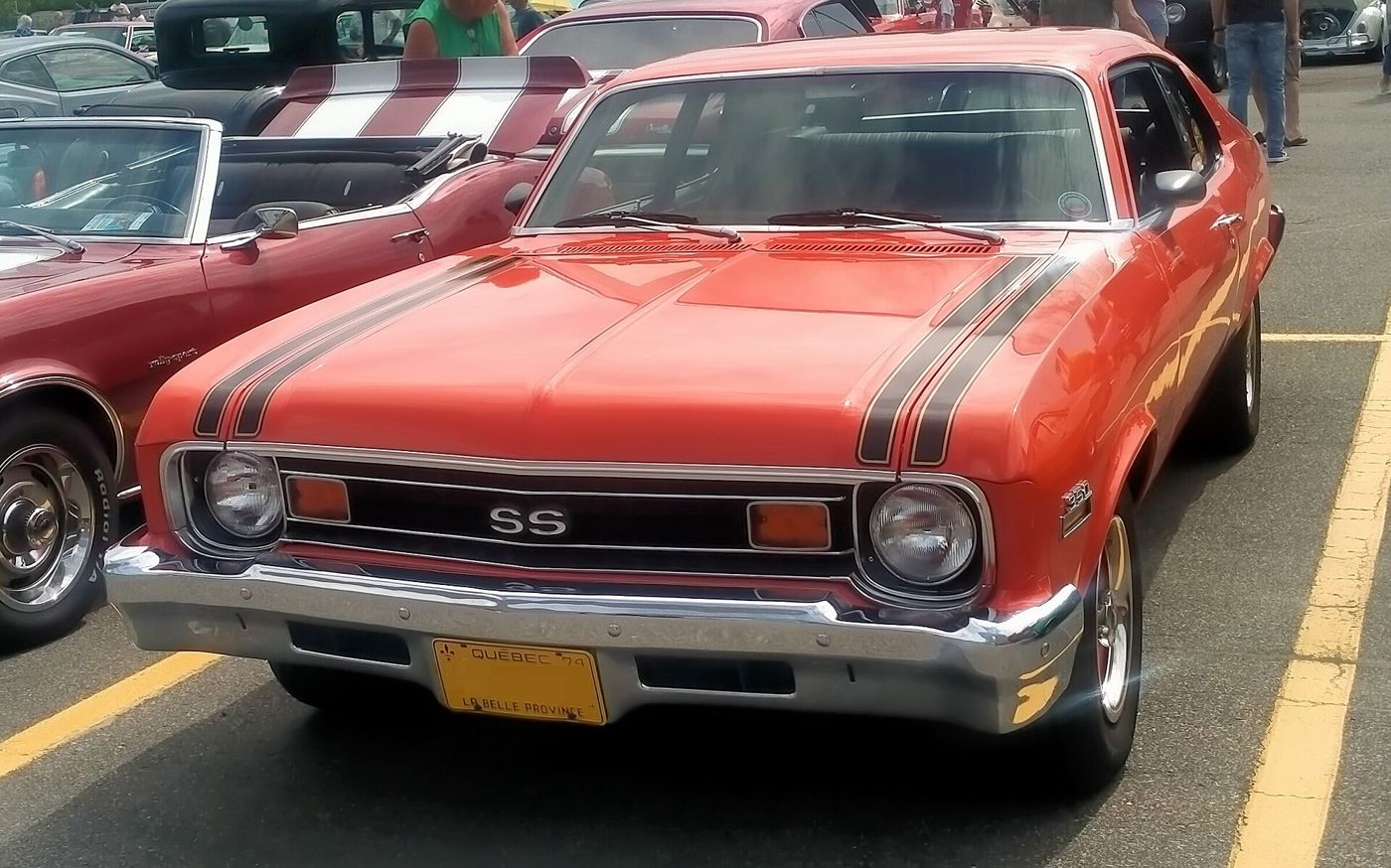 Orange 1974 Chevrolet Nova SS coupe at an outdoor car show, front view, with the SS grille badge, black hood stripes and 350 fender badge