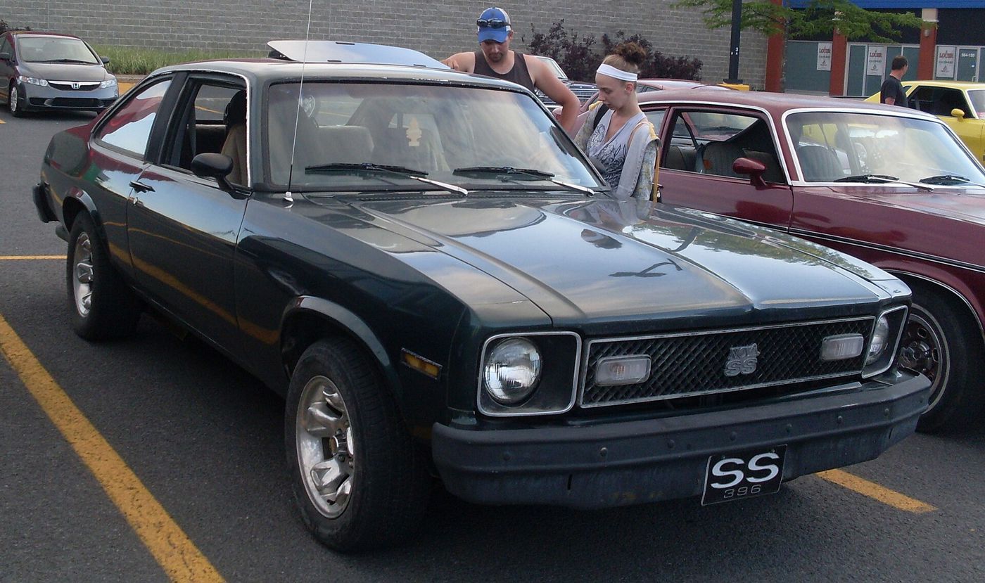 Dark green 1976 Chevrolet Nova SS at an outdoor car show, front three-quarter, showing the black-out grille with Nova SS badge and the SS-style bumper; two visitors stand beside it