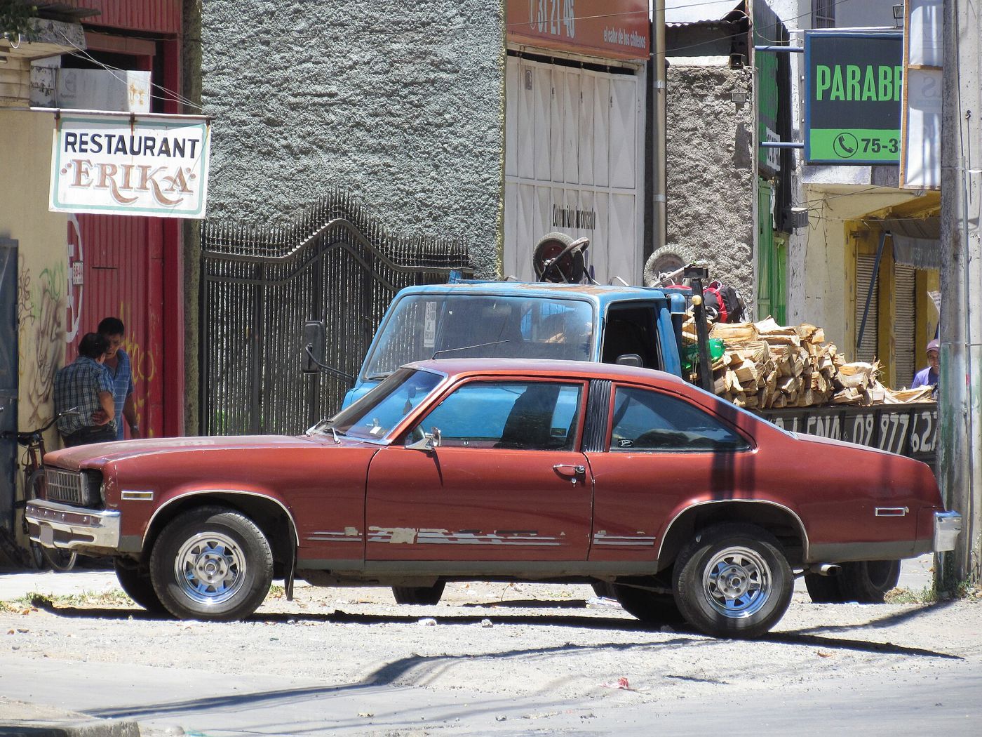 Weathered red 1977 Chevrolet Nova two-door coupe parked on a street in Chile, side view, showing the Nova coupe roofline, rear quarter window and aftermarket wheels