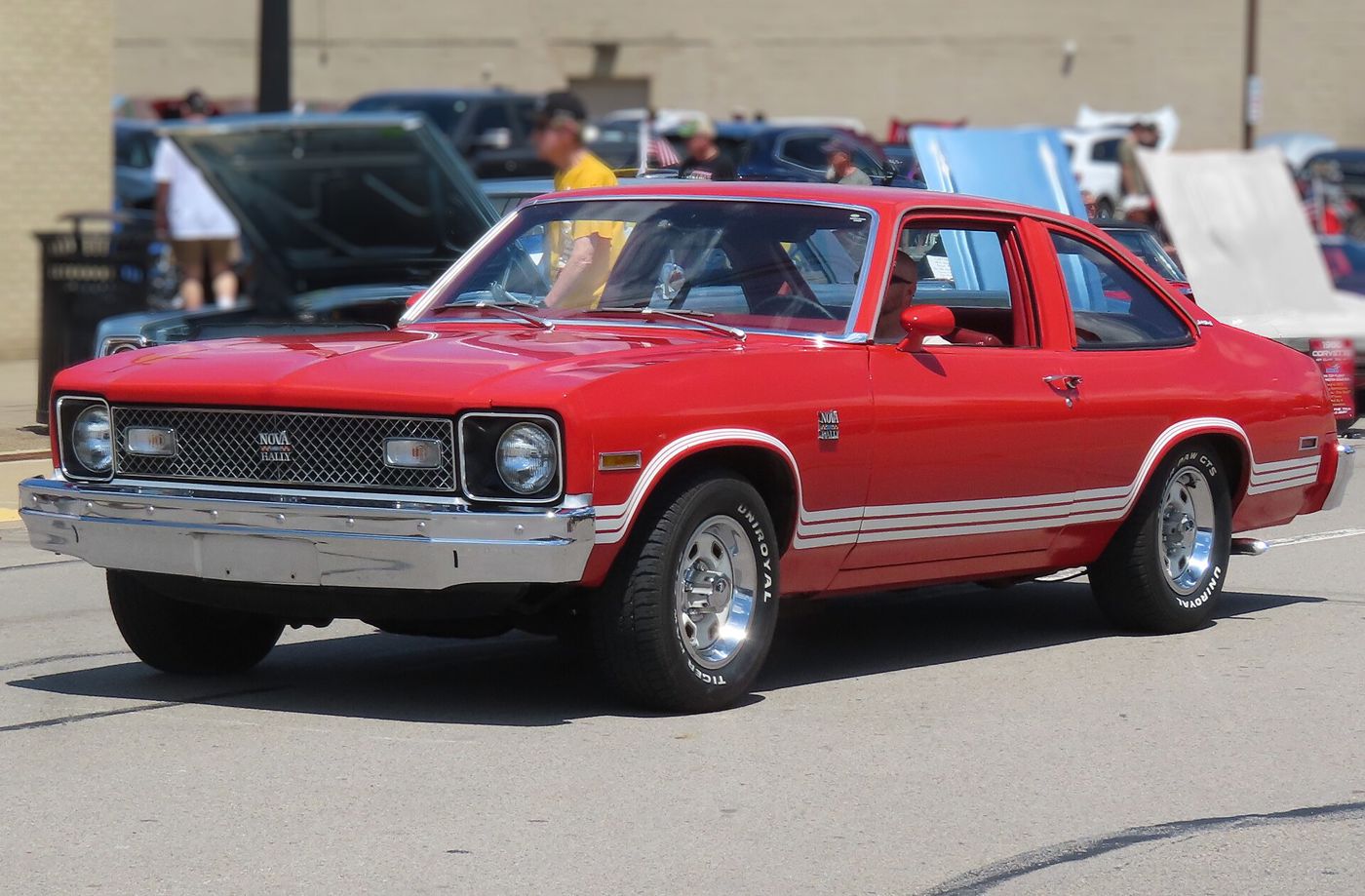 Red 1978 Chevrolet Nova Rally hatchback coupe with white Rally stripes and Nova Rally grille badge, driving slowly at a car show, front three-quarter