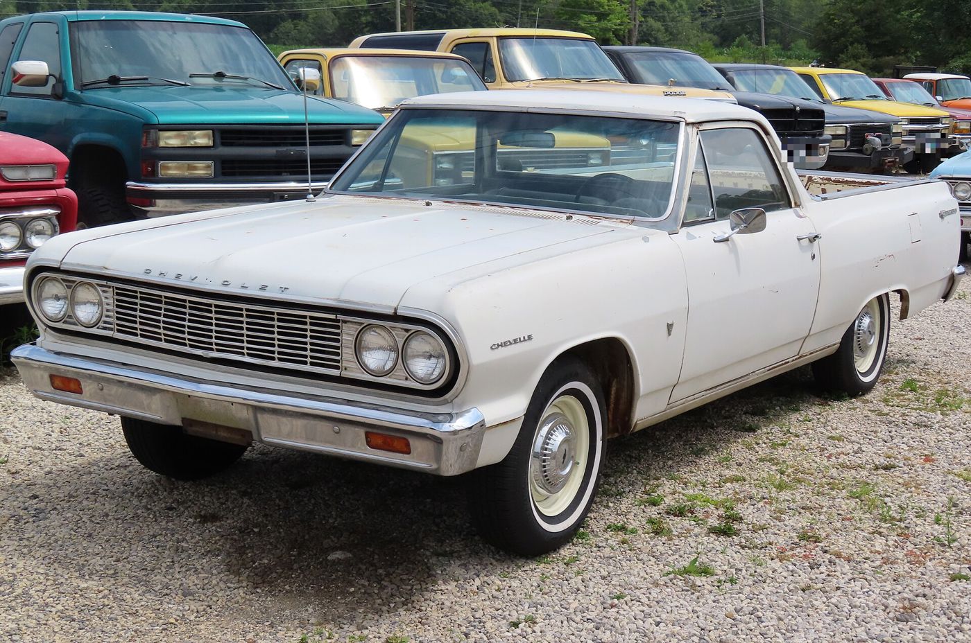 Unrestored white 1964 Chevrolet El Camino on gravel among old pickups, front three-quarter, showing the quad headlamps, horizontal-bar grille and Chevelle fender script