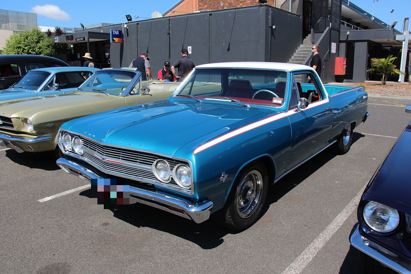 Blue 1965 Chevrolet El Camino with a white roof and white side spear stripe at an Australian car show, front three-quarter, showing the 1965 grille, quad headlamps and crossed-flags V8 fender emblem