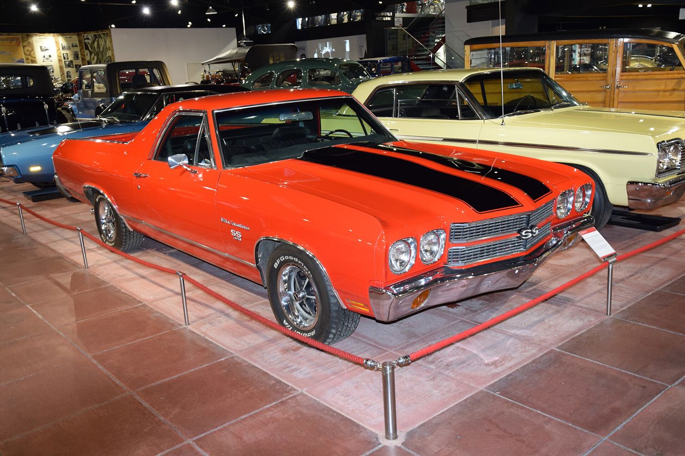 Orange 1970 Chevrolet El Camino SS in a museum hall behind a rope barrier, front three-quarter, with black hood stripes, SS grille badge, El Camino SS fender script and Rally wheels