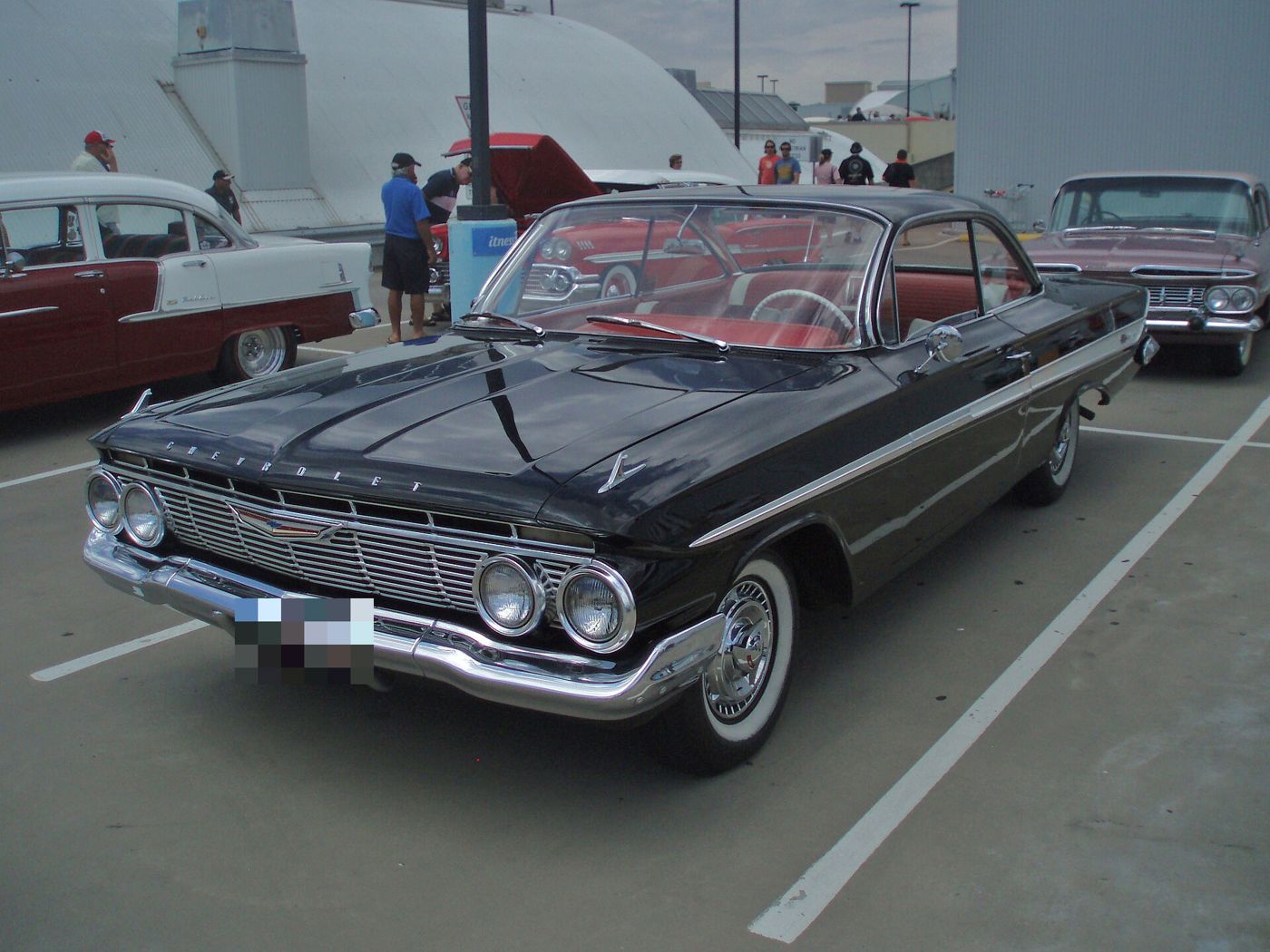 Black 1961 Chevrolet Impala two-door sport coupe with red interior, seen from the front three-quarter at a car show, showing the quad headlamps and full-width grille