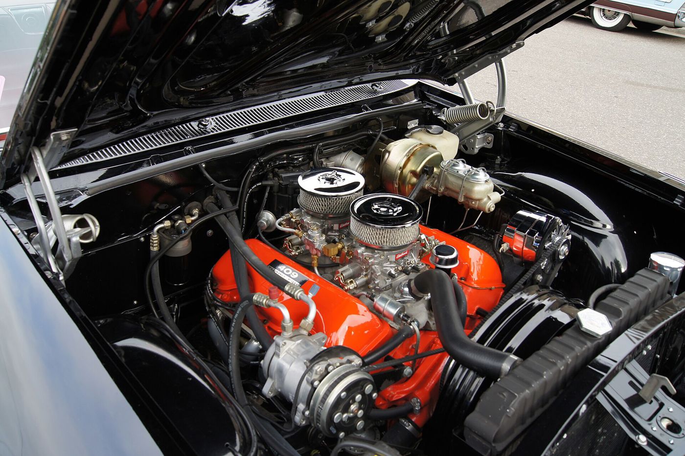 Orange Chevrolet 409 V8 with a 409 Turbo-Fire decal, twin chrome-lidded air cleaners over two Edelbrock carburetors, in the black engine bay of a 1962 Impala SS