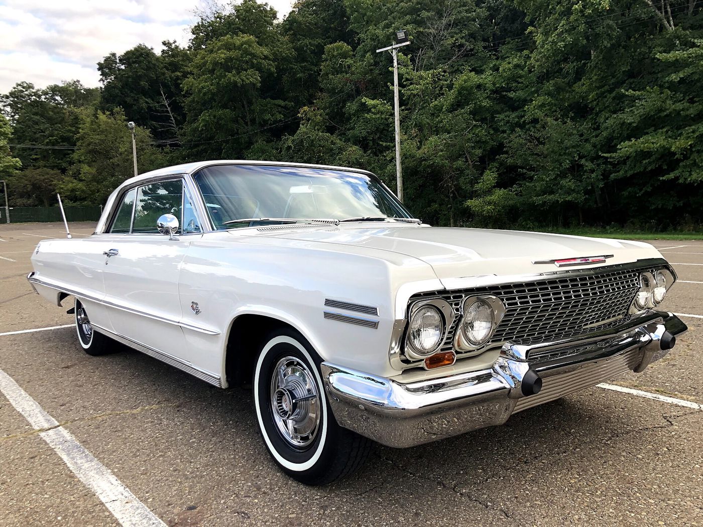 White 1963 Chevrolet Impala two-door hardtop seen from the front three-quarter in an empty parking lot, showing the 1963 grille with its quad headlamps