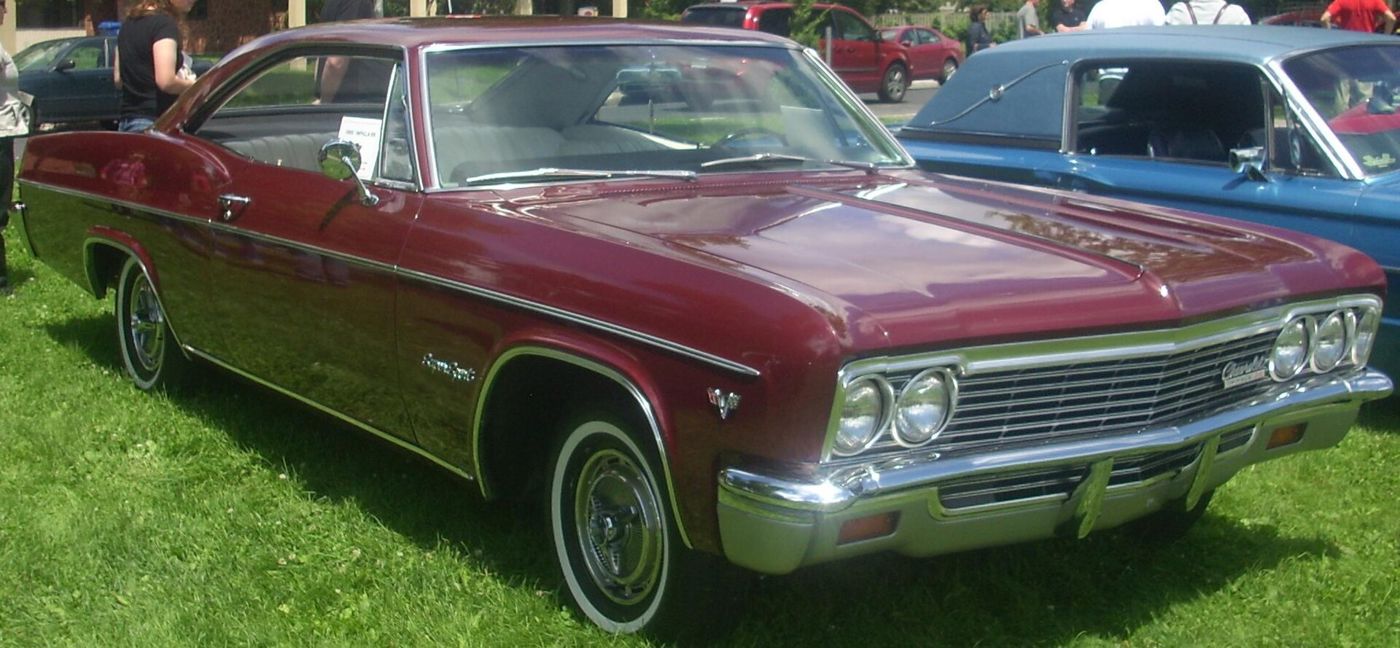 Maroon 1966 Chevrolet Impala SS two-door hardtop on grass at a car show, front three-quarter, with Super Sport script and crossed-flags emblem on the front fender