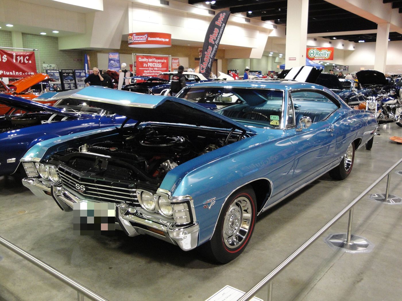 Blue 1967 Chevrolet Impala SS 427 fastback hardtop with its hood raised at an indoor car show, front three-quarter, showing the SS 427 grille badge, crossed-flags fender emblem and Rally wheels with red-stripe tires
