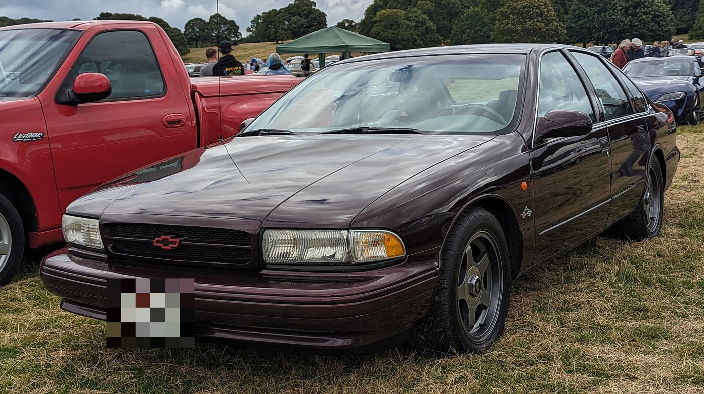 Dark Cherry Metallic 1995 Chevrolet Impala SS four-door sedan at an outdoor car show, front three-quarter, with red-outlined bowtie and Impala emblem on the rear pillar