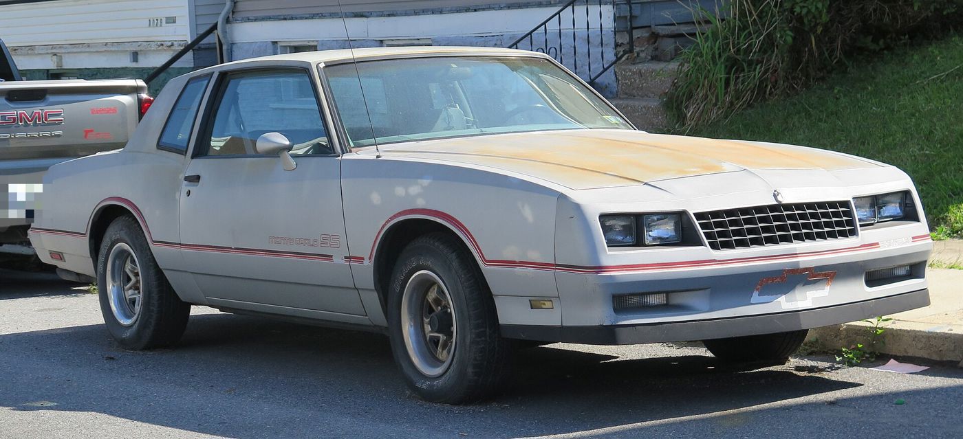 Weathered Silver Metallic 1985 Chevrolet Monte Carlo SS parked at a curb, front three-quarter, with red pinstripes, Monte Carlo SS door lettering and a faded hood