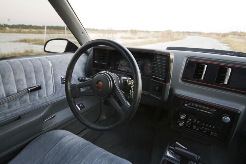 Driver-side view of a 1986 Chevrolet Monte Carlo SS interior with grey cloth seats, the four-spoke steering wheel, gauge cluster and console shifter