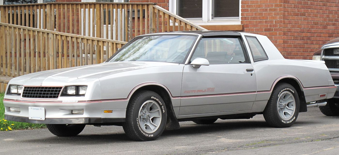 Silver 1986 Chevrolet Monte Carlo SS with a black T-top roof, parked in a driveway, front three-quarter from the driver side, with red pinstripes and Monte Carlo SS door lettering