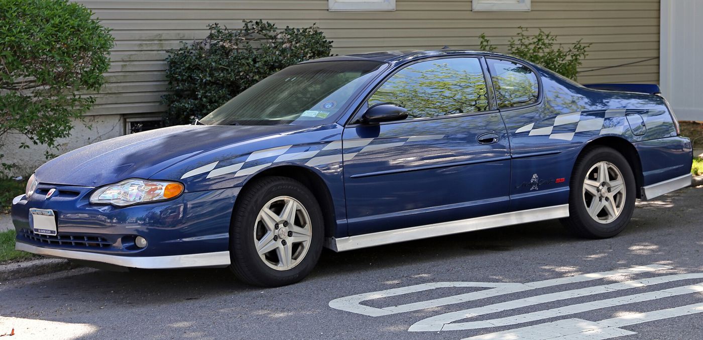 Blue 2003 Chevrolet Monte Carlo SS with silver checkered-flag graphics along the sides and silver lower trim, parked at a curb, side view from the front three-quarter