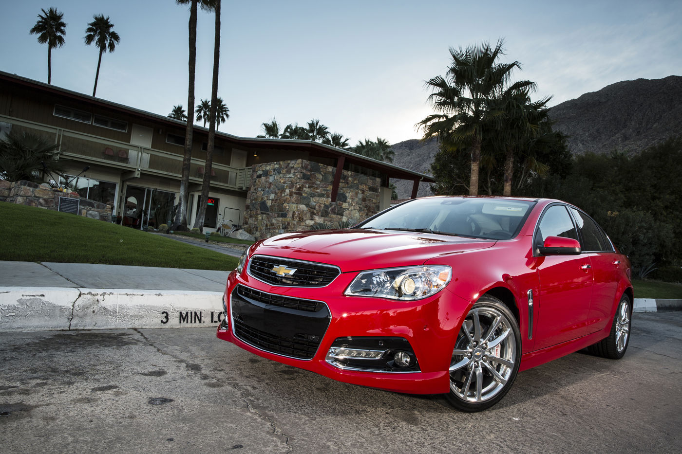 Red 2014 Chevrolet SS four-door sedan, low front three-quarter view in front of a mid-century motel with palm trees and mountains, showing the first-year front fascia and polished wheels