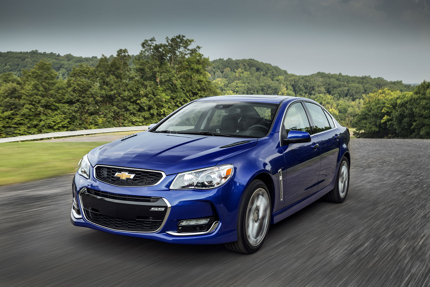 Blue 2016 Chevrolet SS sedan driving on a road lined with trees under a dark sky, front three-quarter, showing the revised 2016 front fascia and hood vents