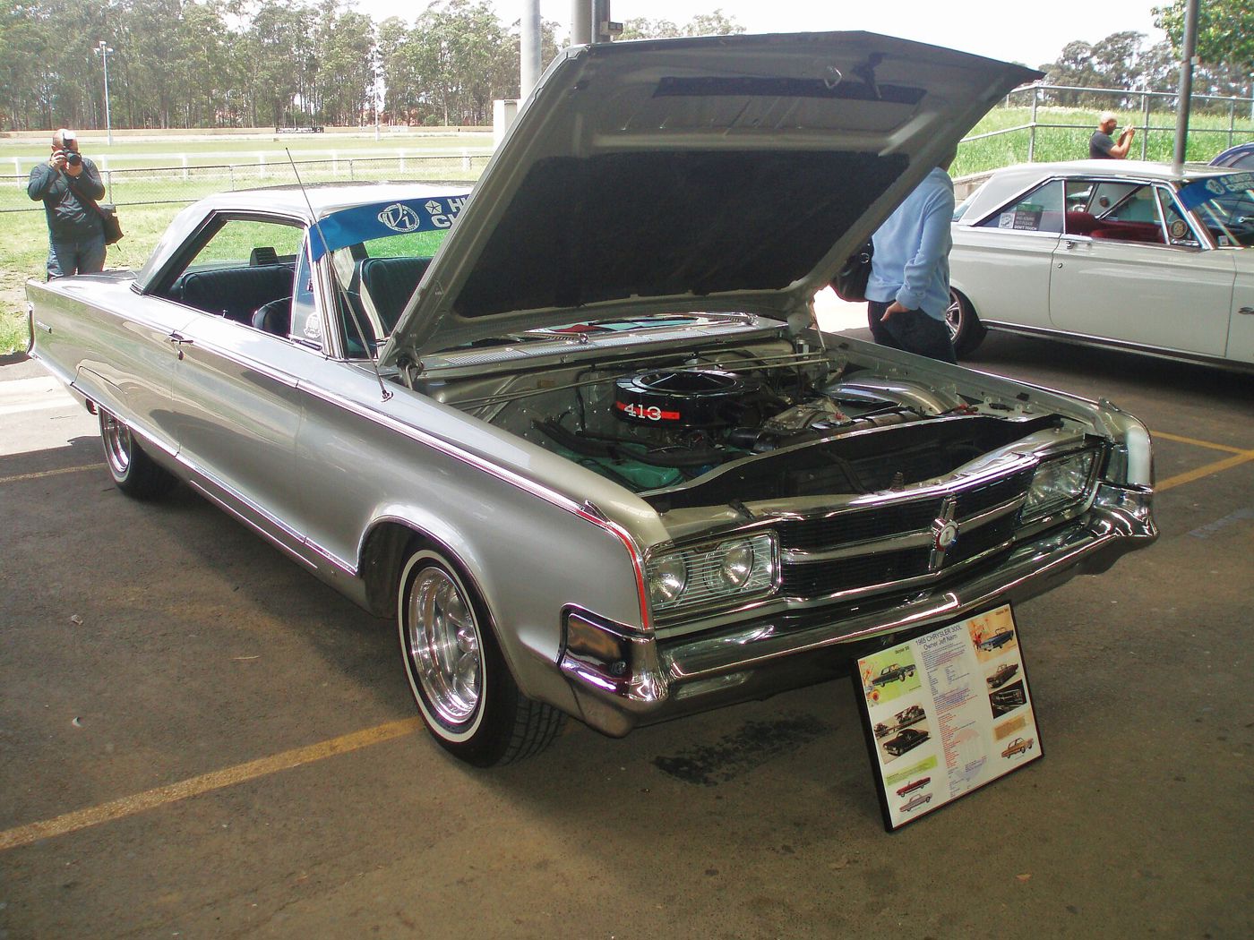 Silver 1965 Chrysler 300L at an indoor show with the hood raised and a display board beside it