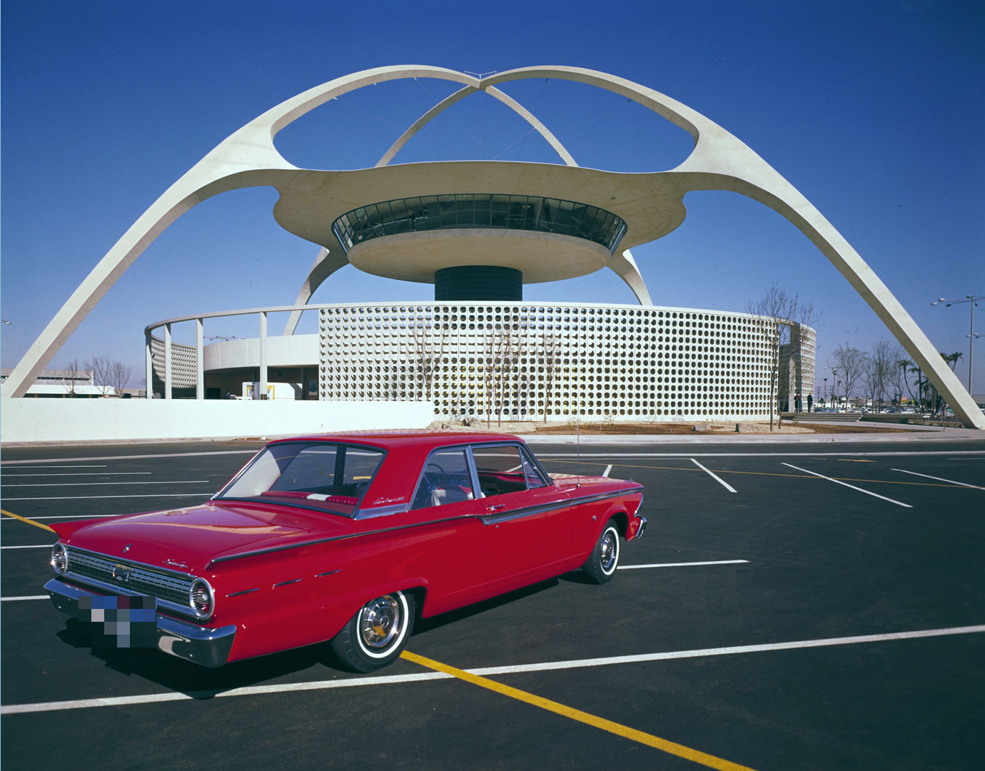 Red 1962 Ford Fairlane 500 two-door sedan seen from the rear three-quarter in a parking lot in front of the arched Theme Building at LAX