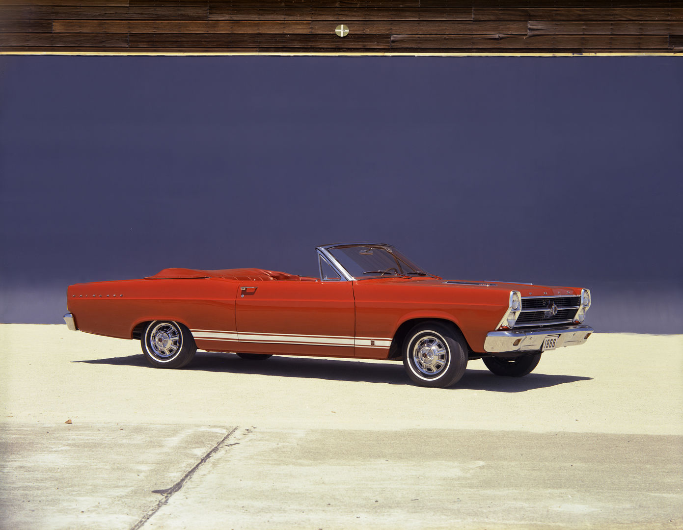 Red 1966 Ford Fairlane GT convertible with the top down, white rocker stripe and hood louvers, parked in front of a dark blue wall