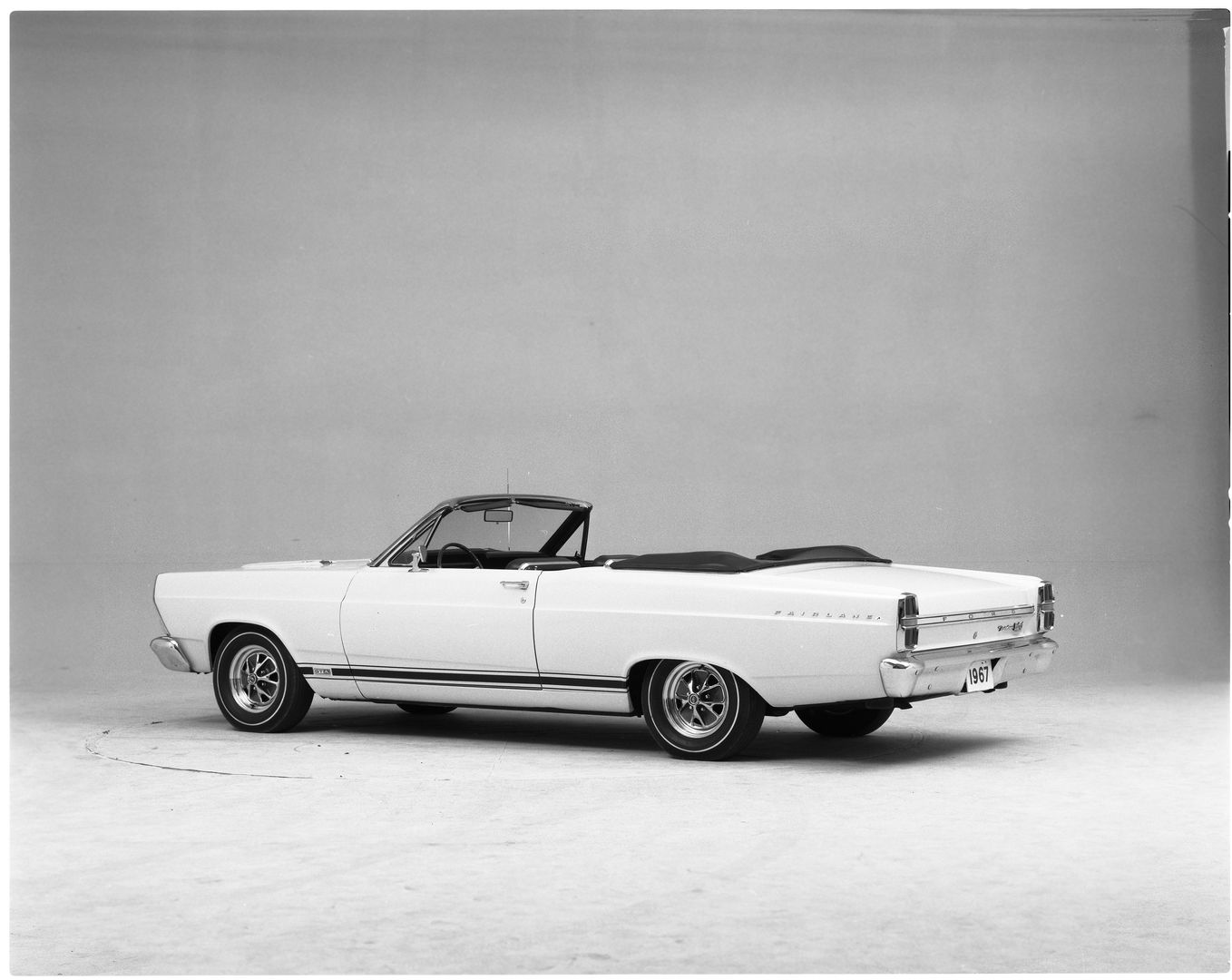 Black-and-white studio photo of a white 1967 Ford Fairlane GTA convertible, top down, from the rear three-quarter, with GTA lettering in the rocker stripe