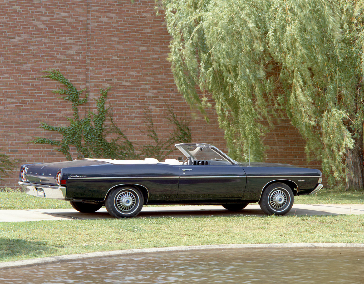Black 1968 Ford Fairlane 500 convertible with the top down, side and rear three-quarter view beside a brick wall and a willow tree