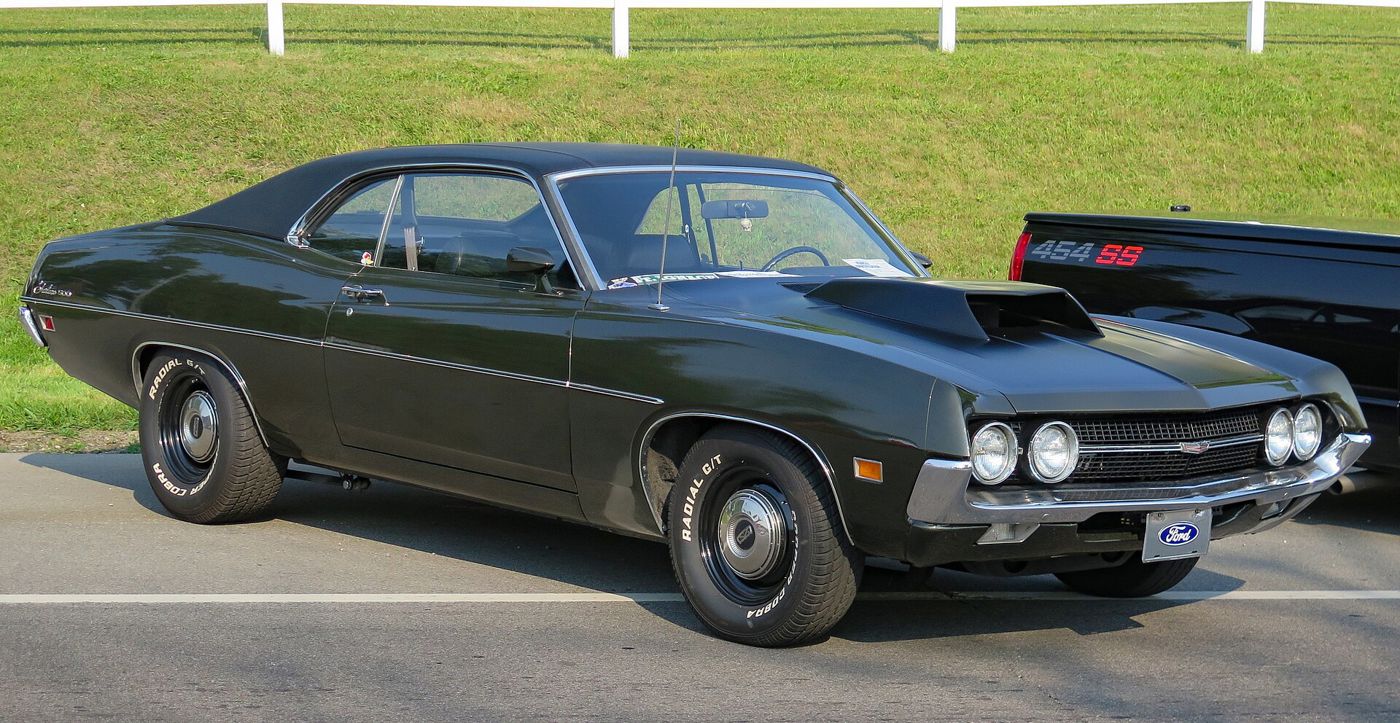 Dark green 1970 Ford Fairlane 500 two-door hardtop with a black vinyl roof, raised hood scoop and wide tires, front three-quarter view at a car show