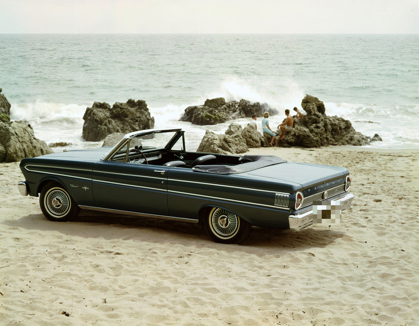 Dark 1964 Ford Falcon Sprint convertible seen from the rear three-quarter on sand, with FALCON lettering across the rear panel and Sprint script on the front fender