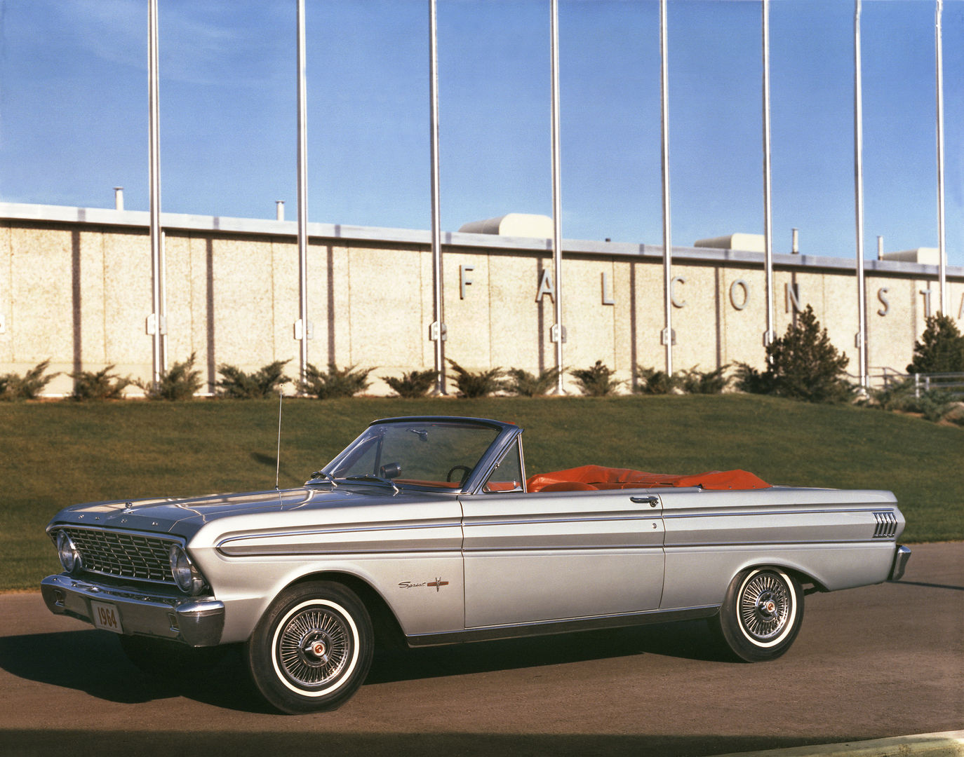 Silver 1964 Ford Falcon Sprint convertible, top down, parked in front of a Ford building with FALCON lettering on the wall
