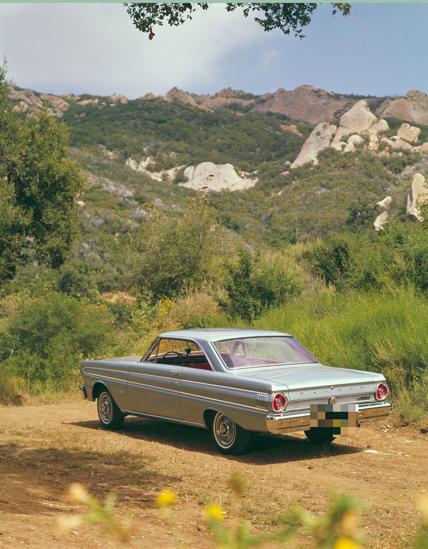 Silver-blue 1964 Ford Falcon Sprint two-door hardtop seen from the rear three-quarter on a dirt track below rocky hills