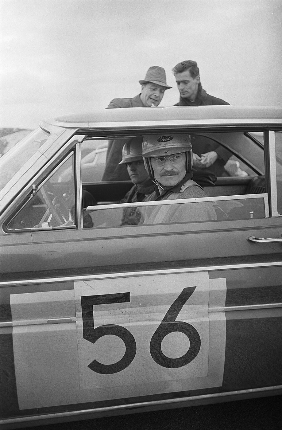 Black-and-white 1964 photo of a rally crew in helmets seated in a Ford Falcon Sprint with competition number 56 on the door, two men leaning on the roof