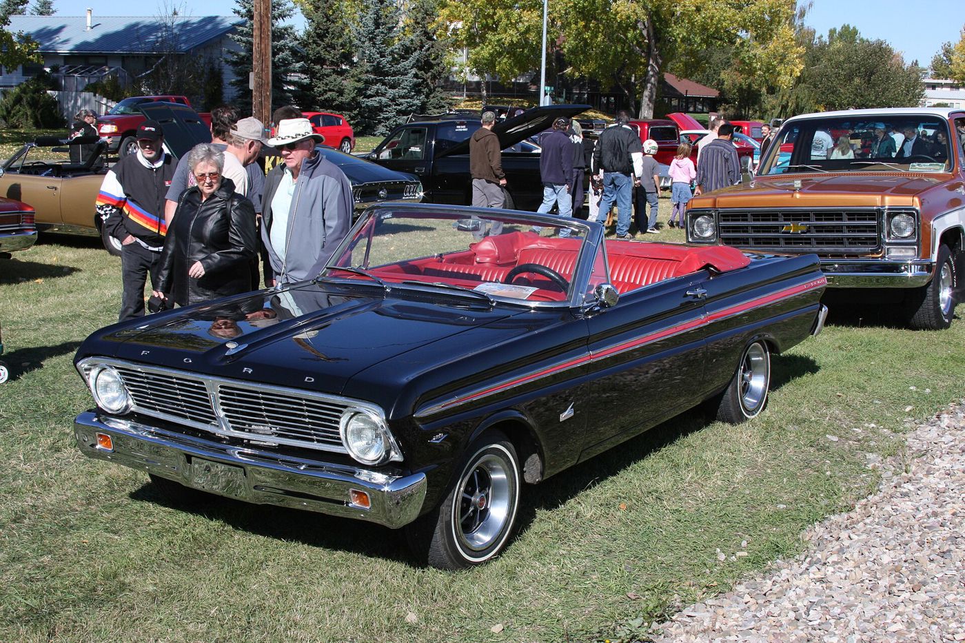 Black 1965 Ford Falcon convertible with red bucket seats and a red side stripe at an outdoor car show, seen from the front three-quarter