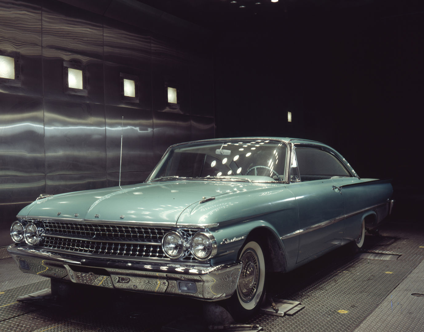Pale turquoise 1961 Ford Galaxie Starliner hardtop on test rollers inside a steel-walled test chamber lit by overhead lamps